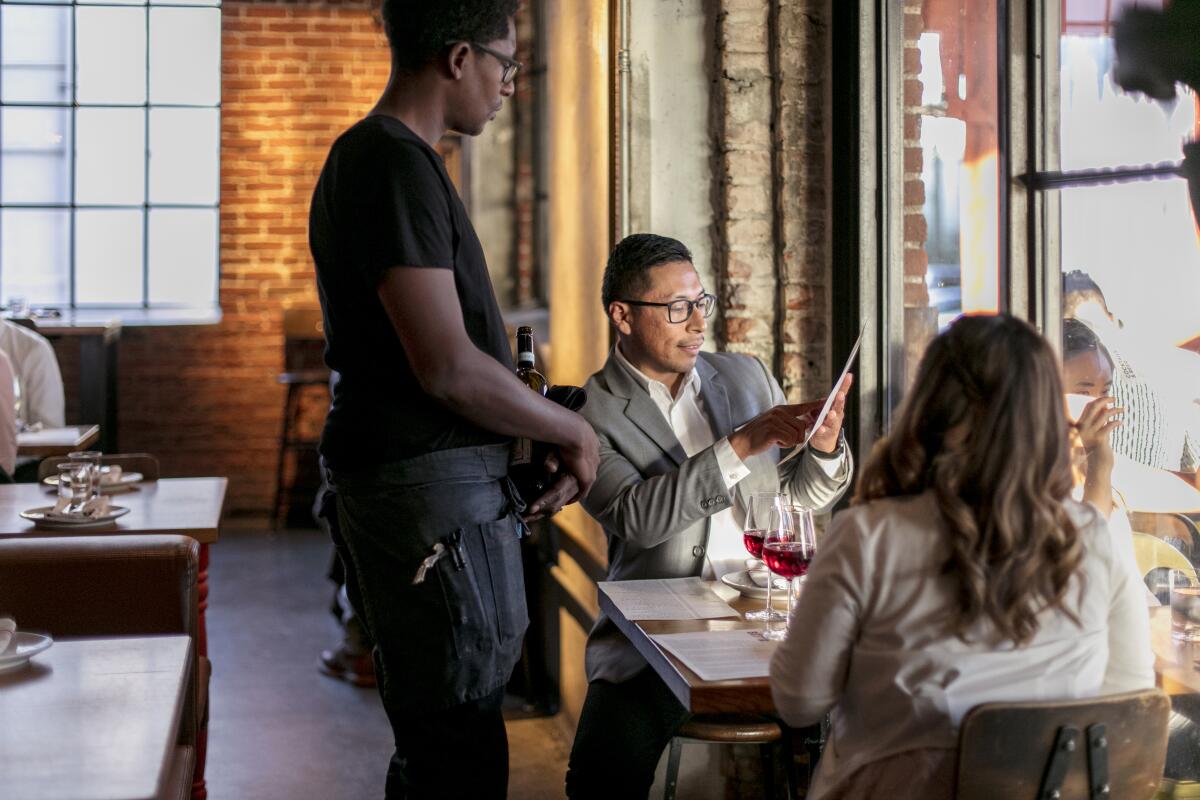 Two seated diners gesture to a menu while their server stands at the table.