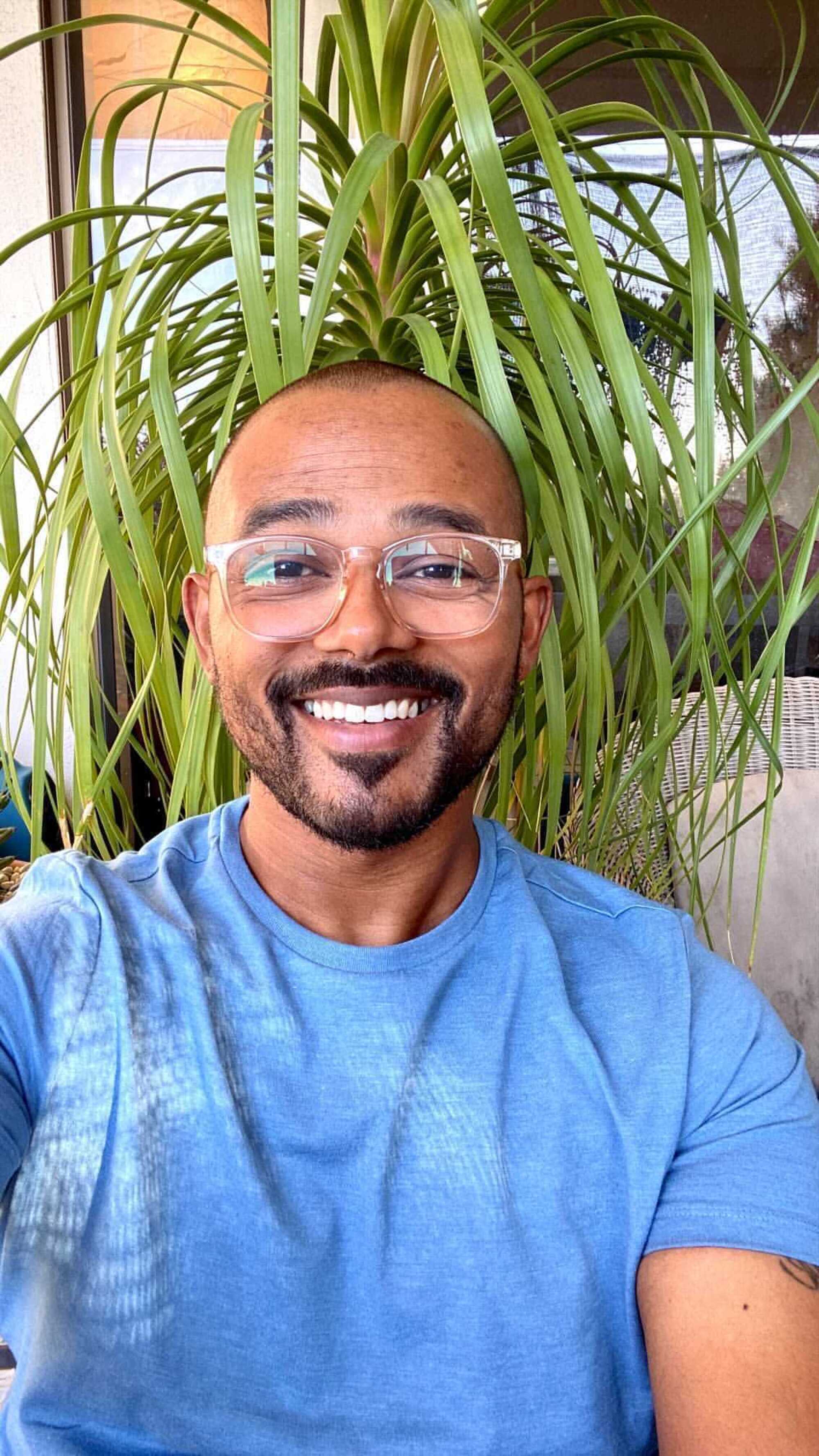 A smiling man wearing clear-framed glasses and a blue T-shirt, in front of a hanging plant.