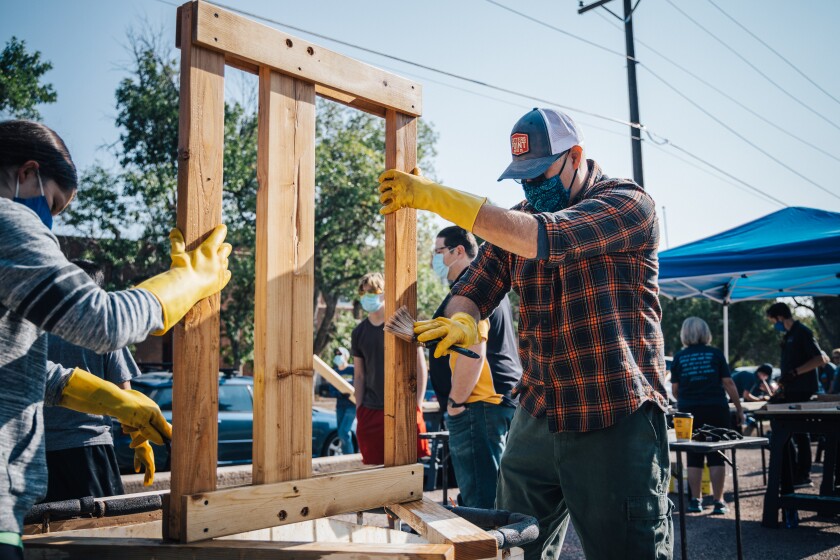 Two people wearing masks and rubber gloves hold a wooden frame upright as they build a bed