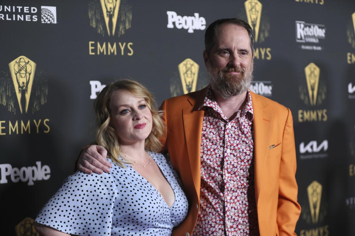 Brendan Hunt in an orange blazer and red patterned shirt with his arm around Shannon Nelson, in a blue dotted dress