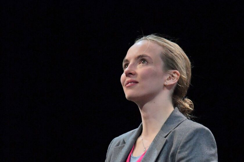 Jodie Comer is standing in a dark stage wearing a grey suit and pink shirt, folding her hands and staring upward