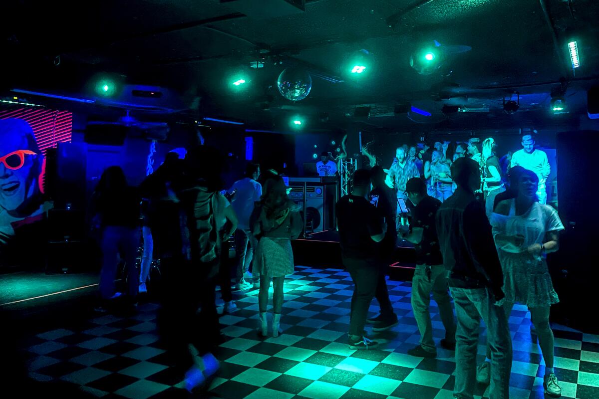People dancing in a dark club under blue and green lights on a checkerboard floor.
