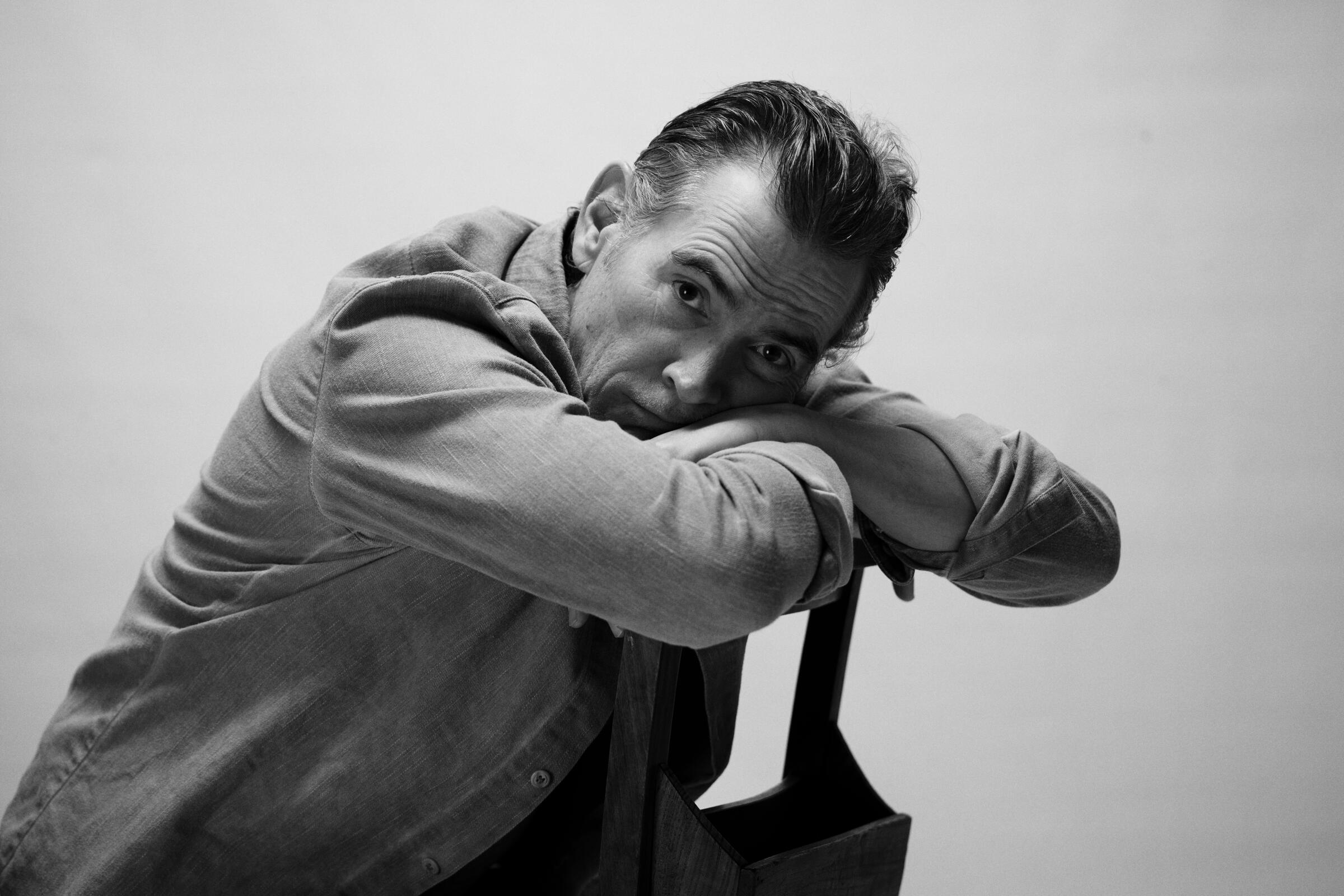 Billy Crudup sits backward in a chair, resting his head and arms on its back, in a black-and-white portrait.