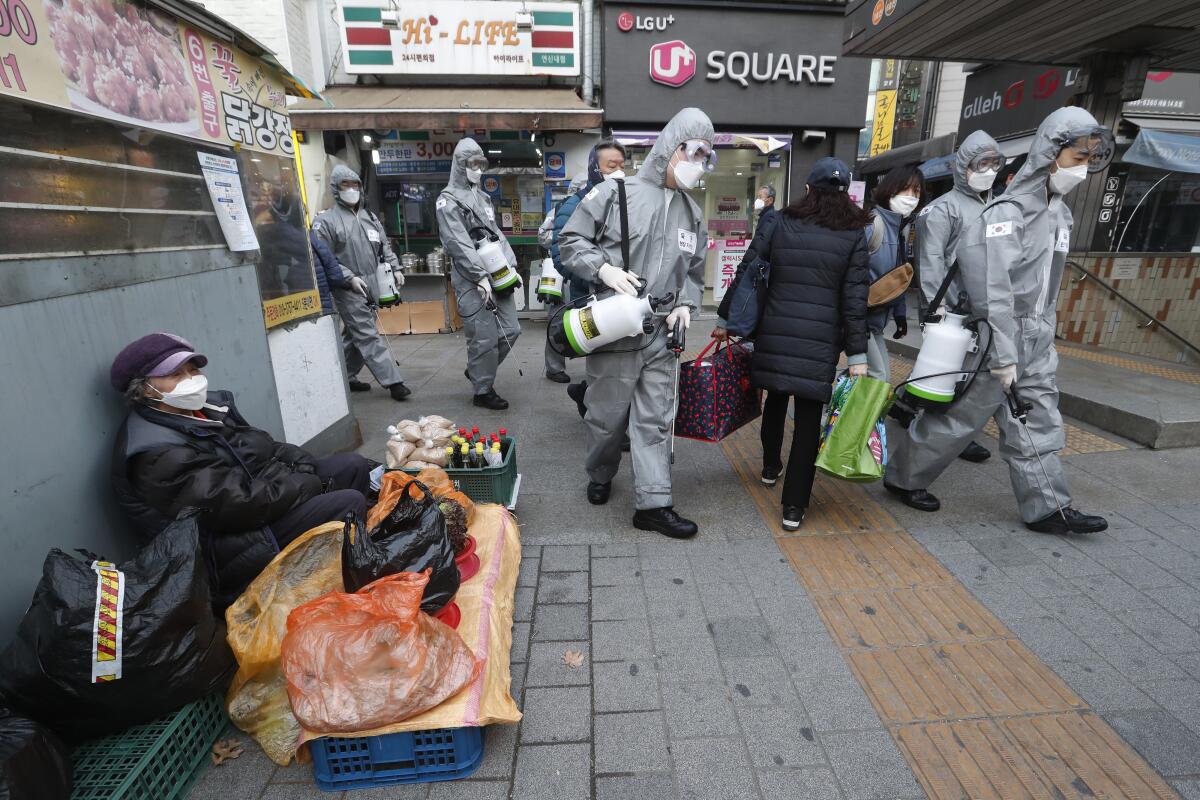 People in protective gear walk by a senior woman sidewalk vendor in Seoul, all wearing face masks.