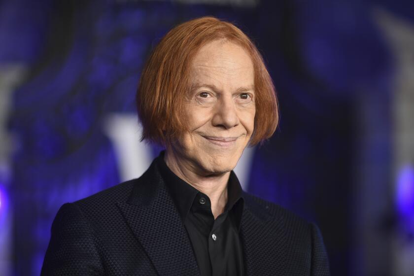 Danny Elfman is posing and smiling with his mouth closed while wearing a dark blue suit with subtle stripes