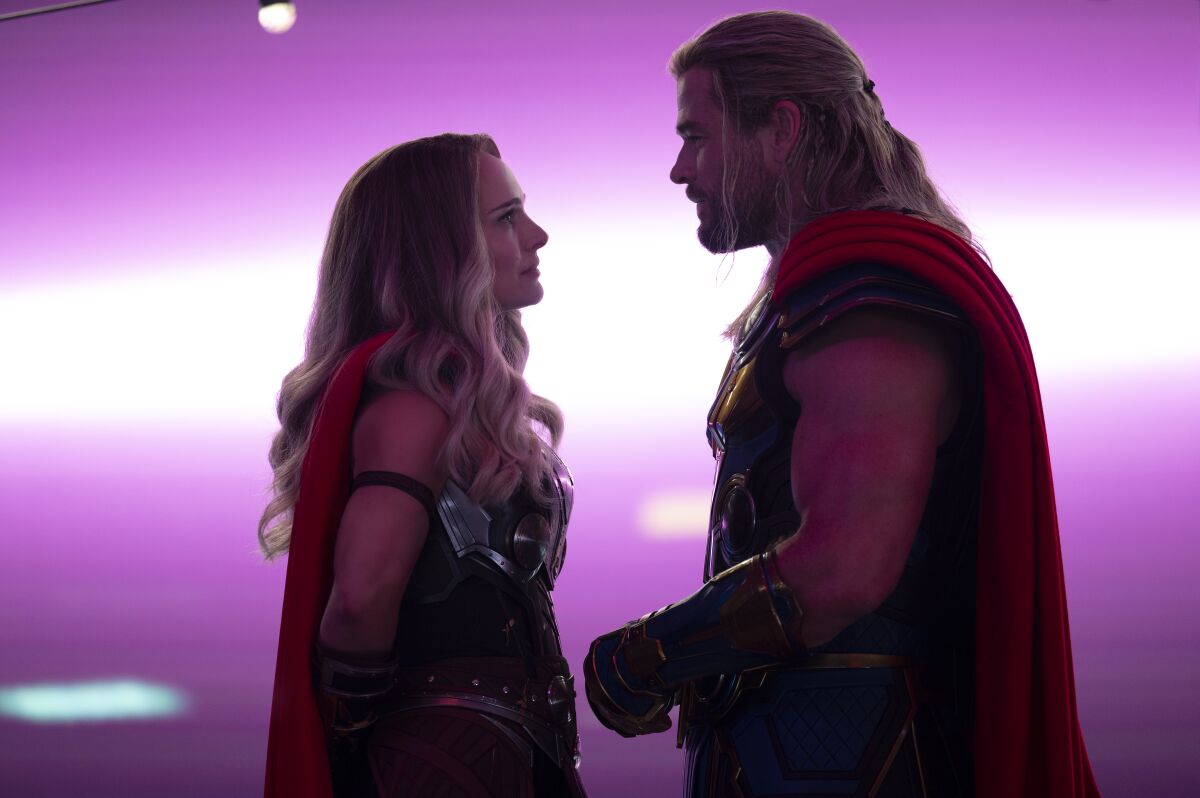 A woman in a superhero costume stands face-to-face with a man in a matching costume under purple lighting.