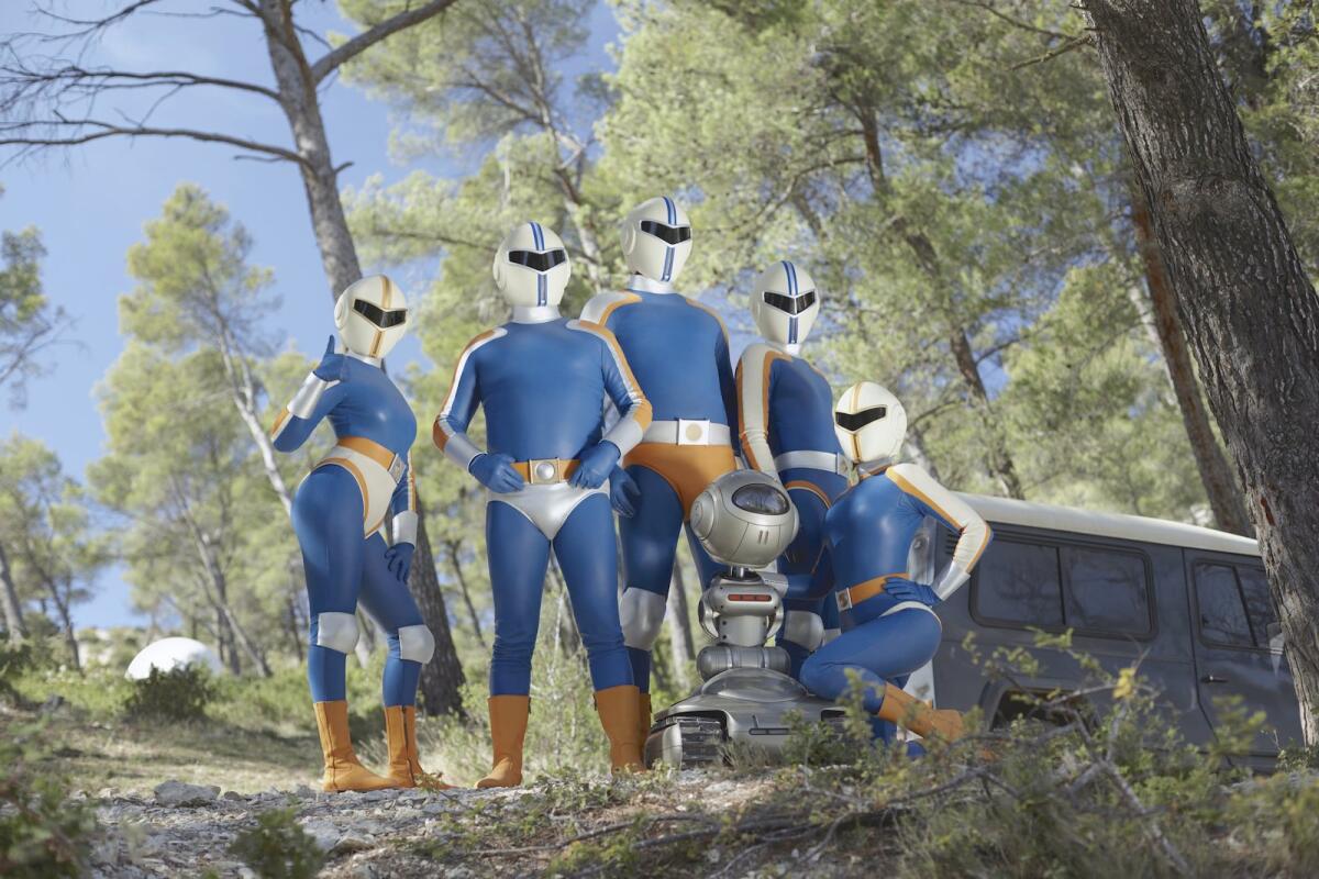 Five costumed superheroes and a robot stand outdoors in the movie "Smoking Causes Coughing."