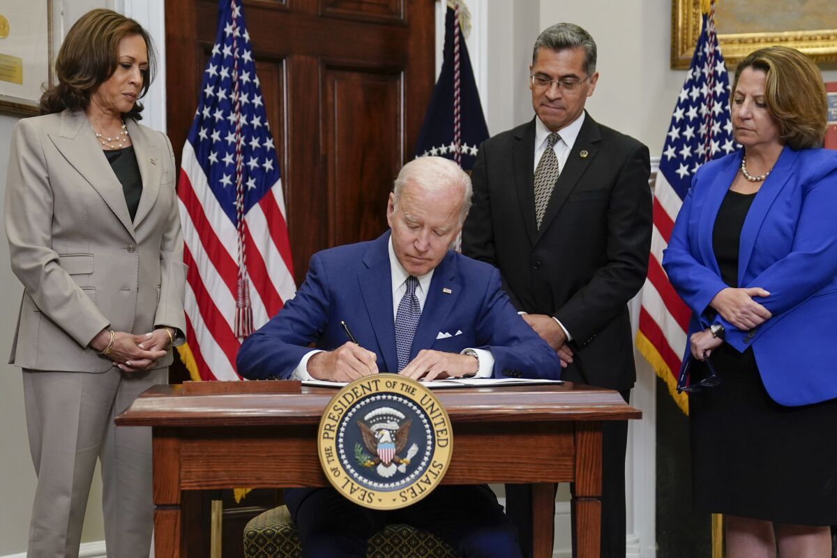President Biden sits at a ceremonial desk with Vice President Kamala Harris and others at his side.