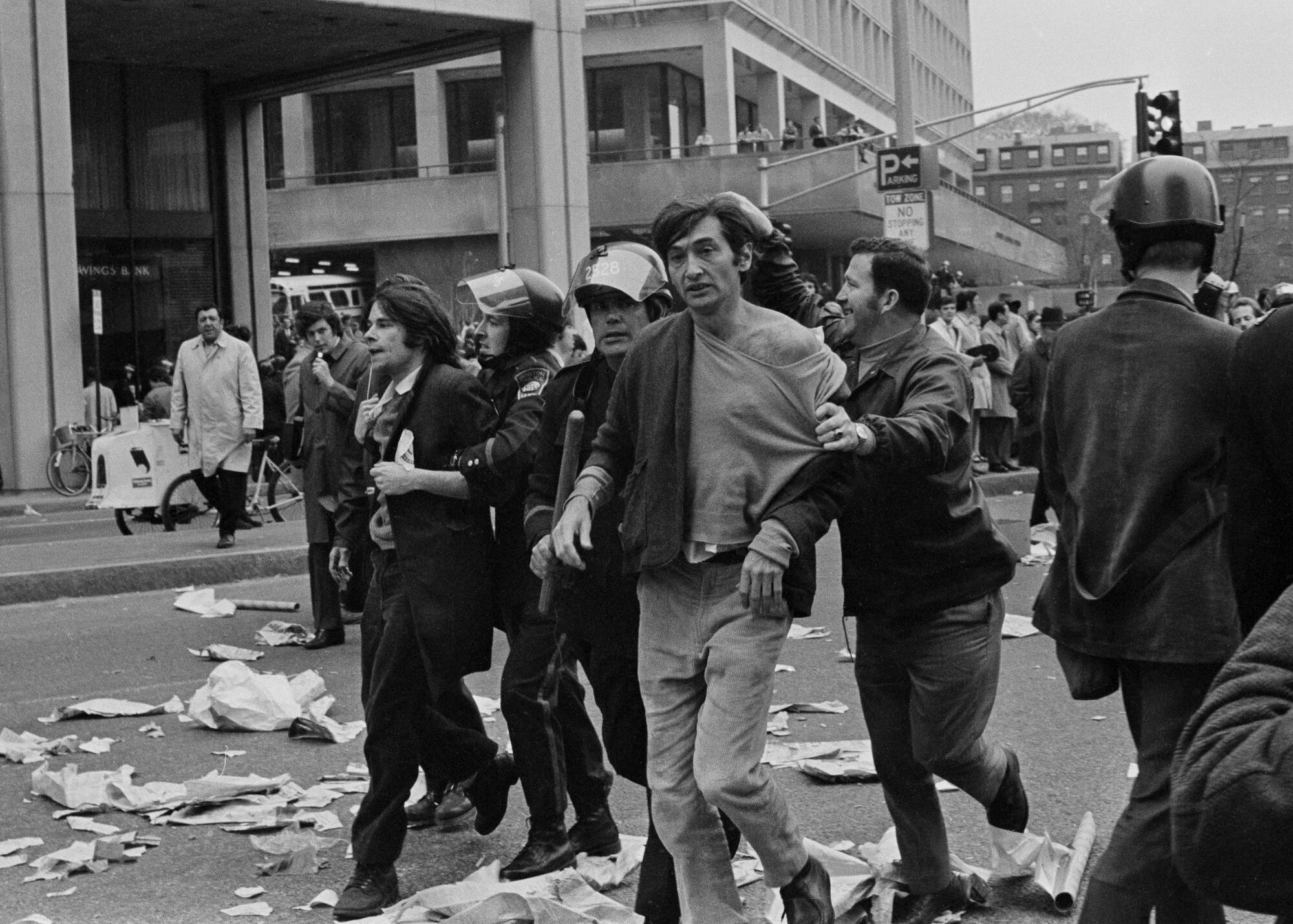 A black-and-white photograph of police leading Howard Zinn and another person away on a street littered with paper