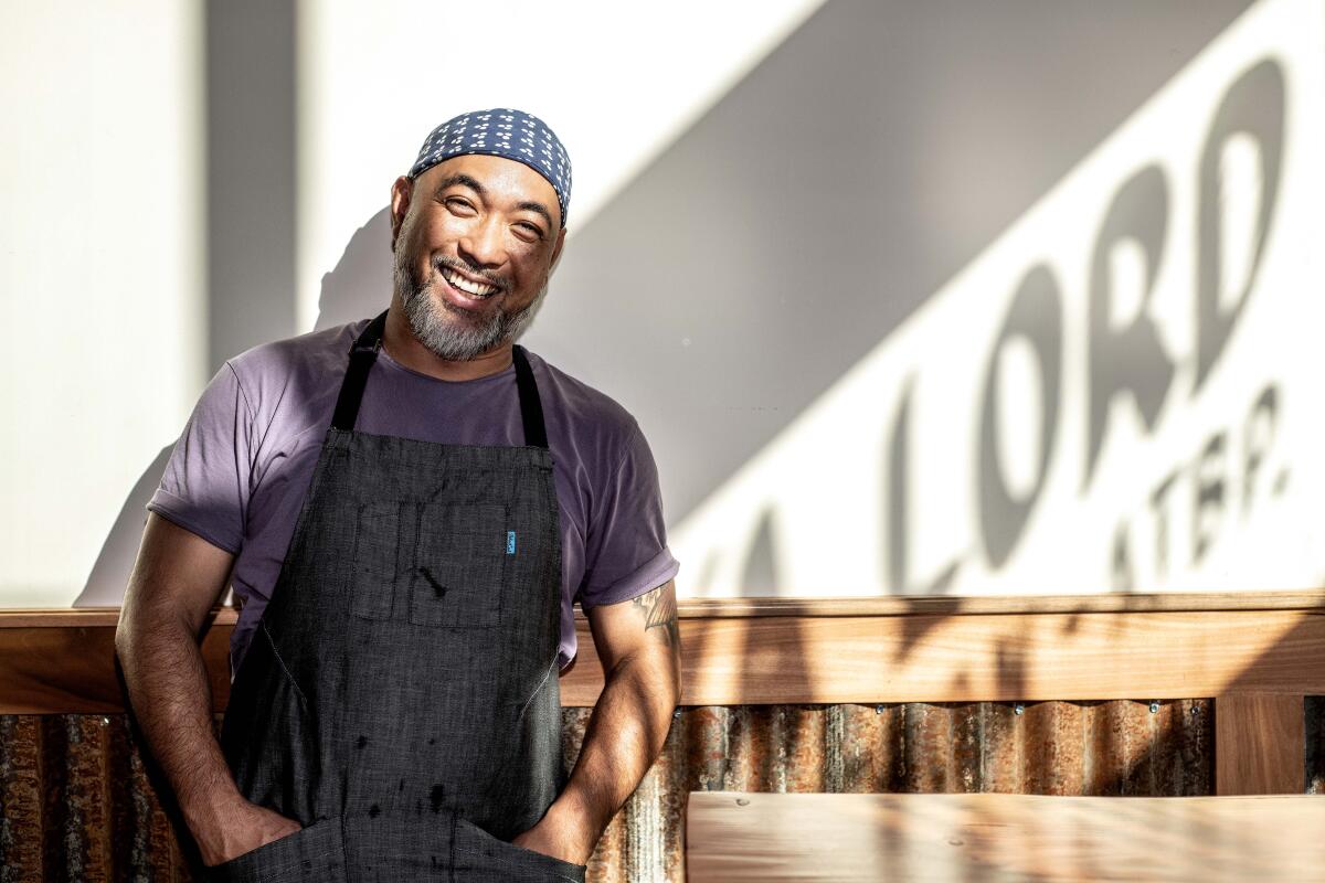 A manin a beanie and an apron smiles for a photo as a he leans against a wall.