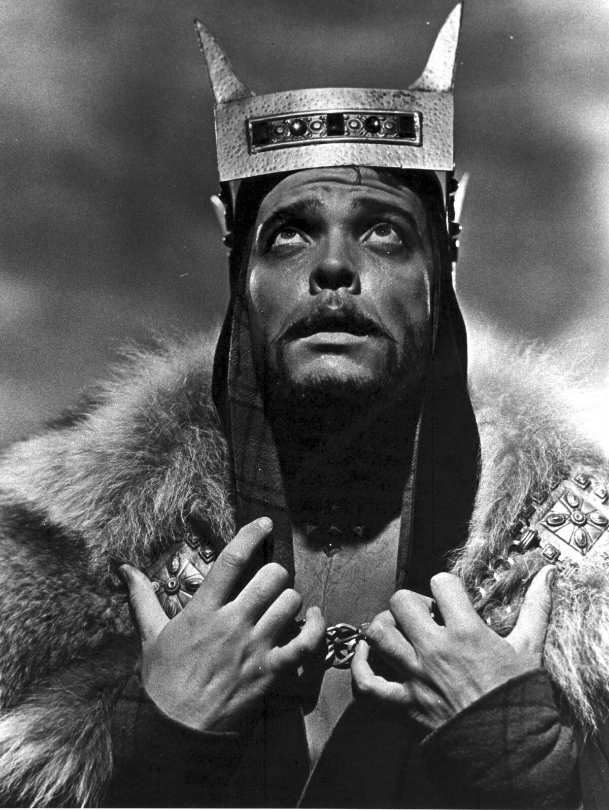 A black-and-white photo of a man dressed as a king looking up and looking distressed 