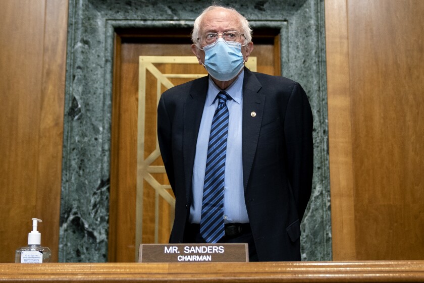 Bernie Sanders stands in a suit and a mask with his hands behind his back in a Senate meeting room