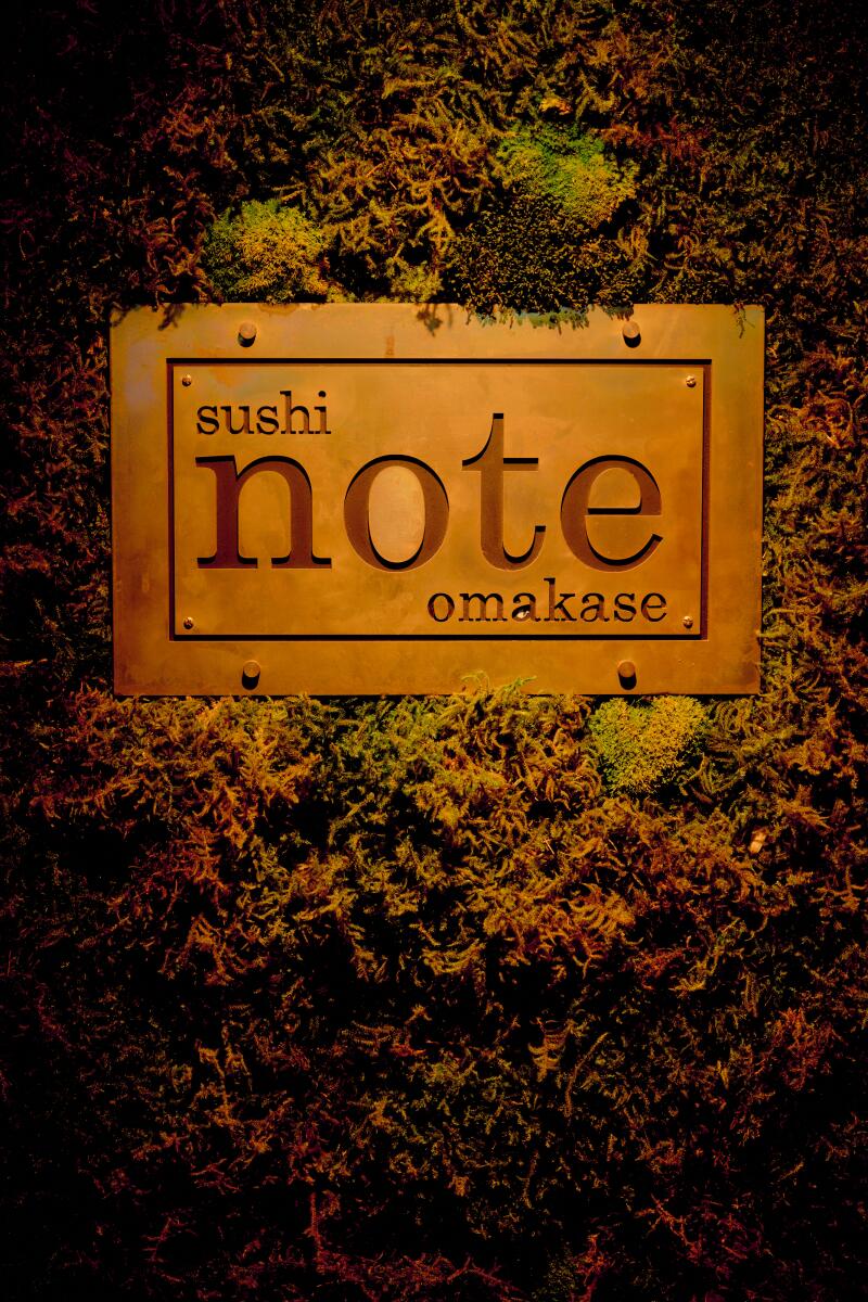 A sign that reads "Sushi Note Omakase" hangs among foliage