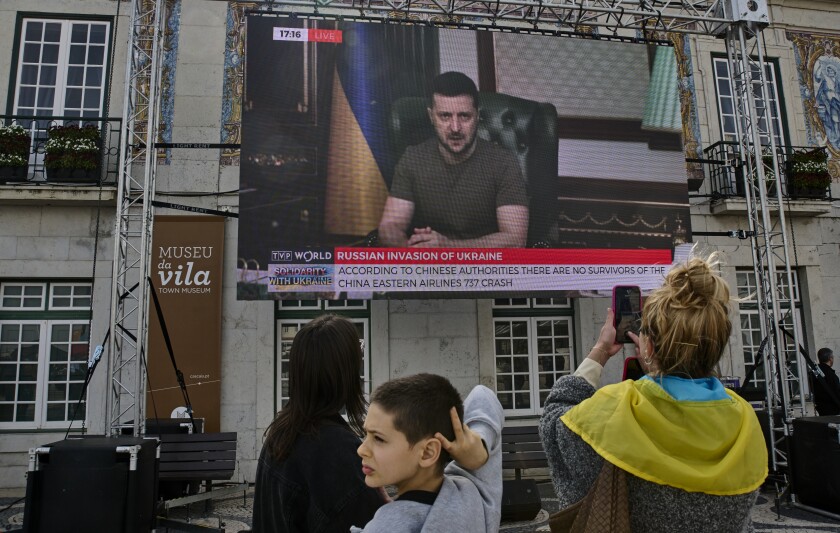 A woman takes a picture of Ukrainian President Volodymyr Zelensky on a giant screen while standing outside of City Hall.