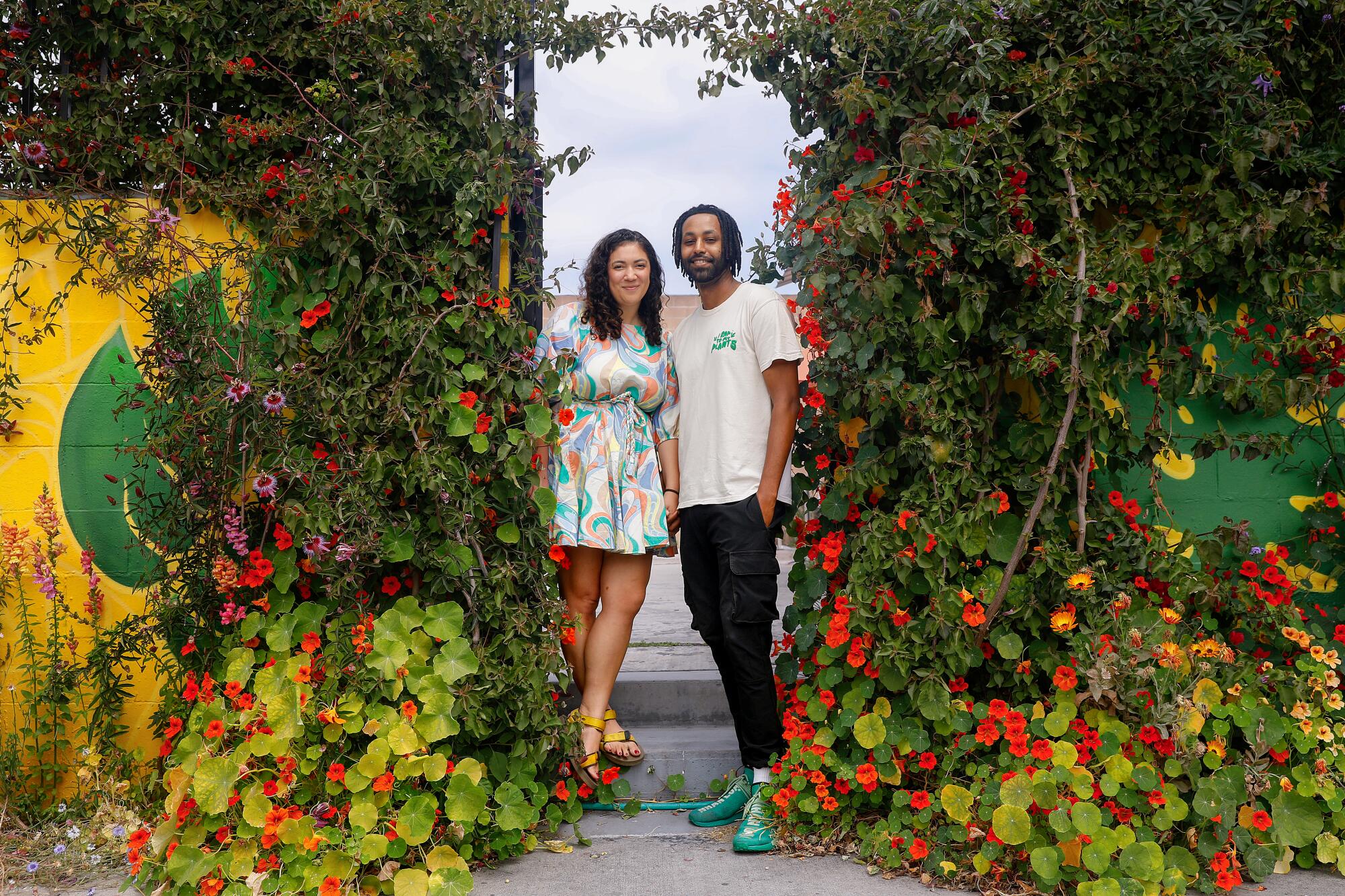 A couple standing in the entrance to their plant shop, surrounded by greenery and flowering plants