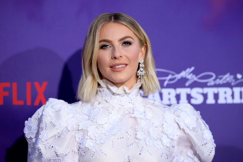 A blond woman poses against a purple background in a puffy white blouse