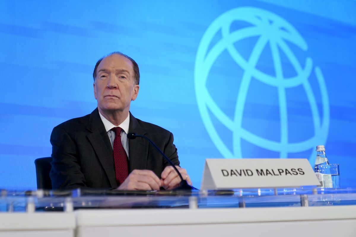 A man in a suit sitting behind a nameplate reading "David Malpass," against a blue background with a stylized globe