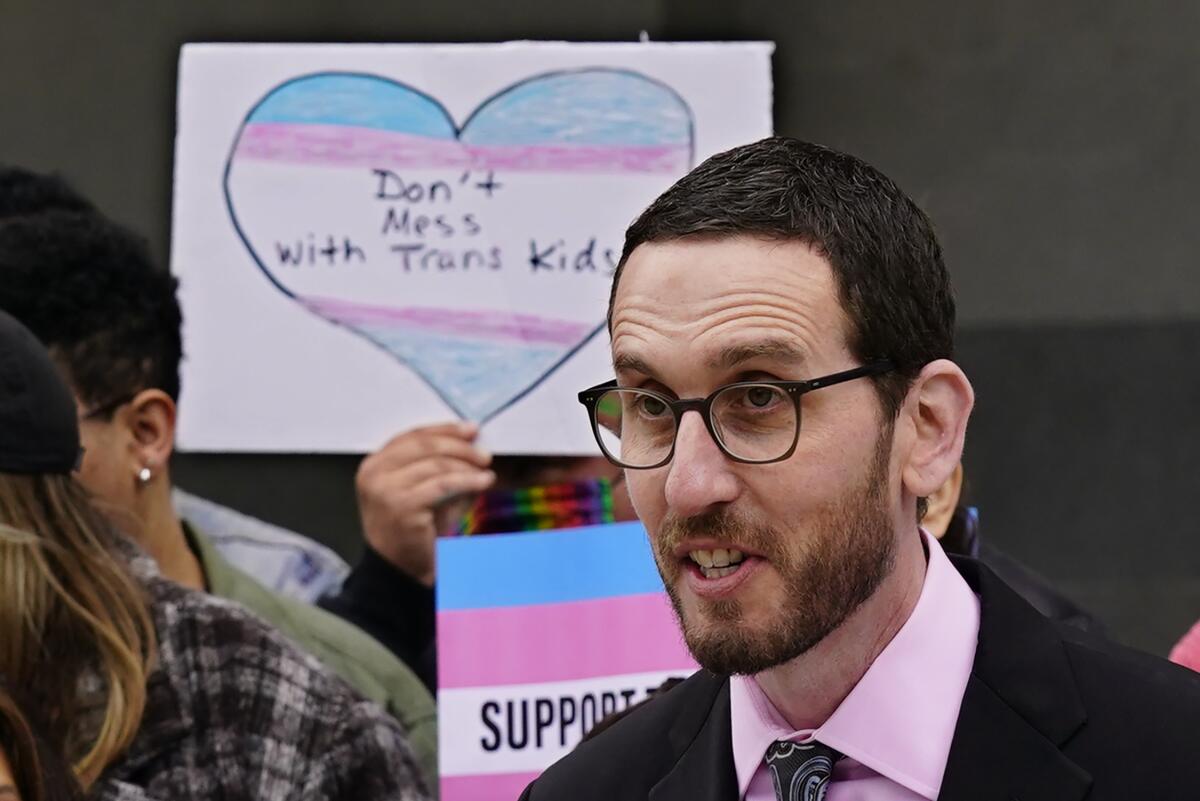 A man speaks as people raise pink-and-blue signs behind him, one with the words "Don't mess with trans kids" inside a heart