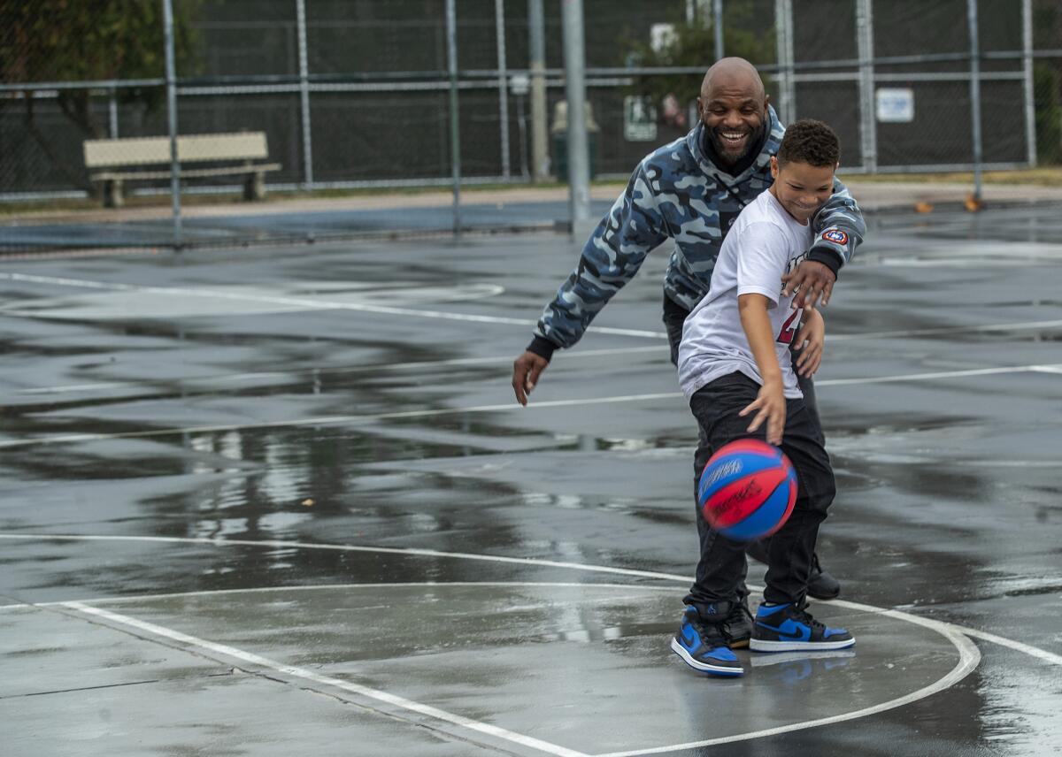 A boy dribbles a basketball on a wet outdoor court as his father guards him