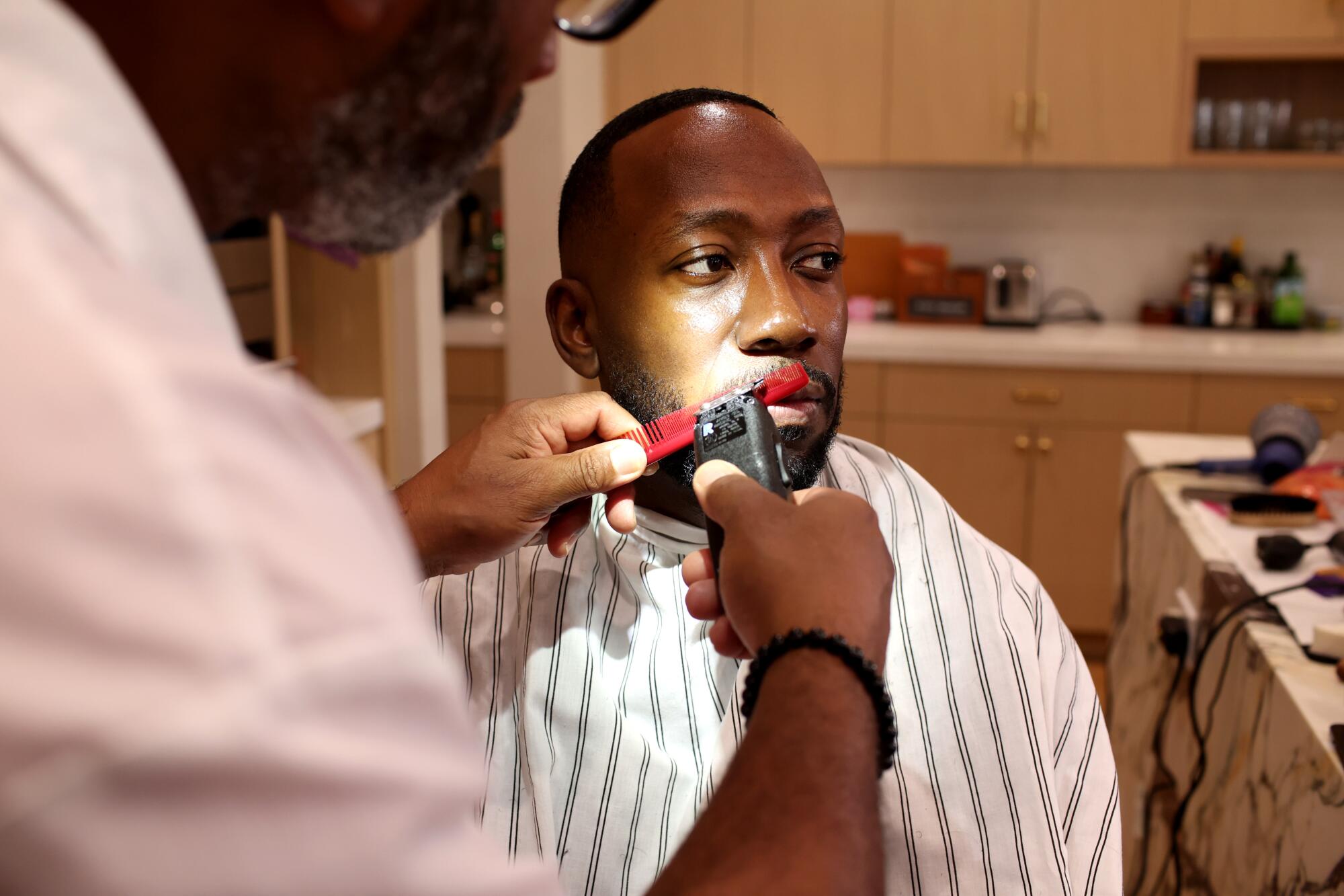 A man holding a clipper and red comb trims the beard of a man sitting and looking to his side.