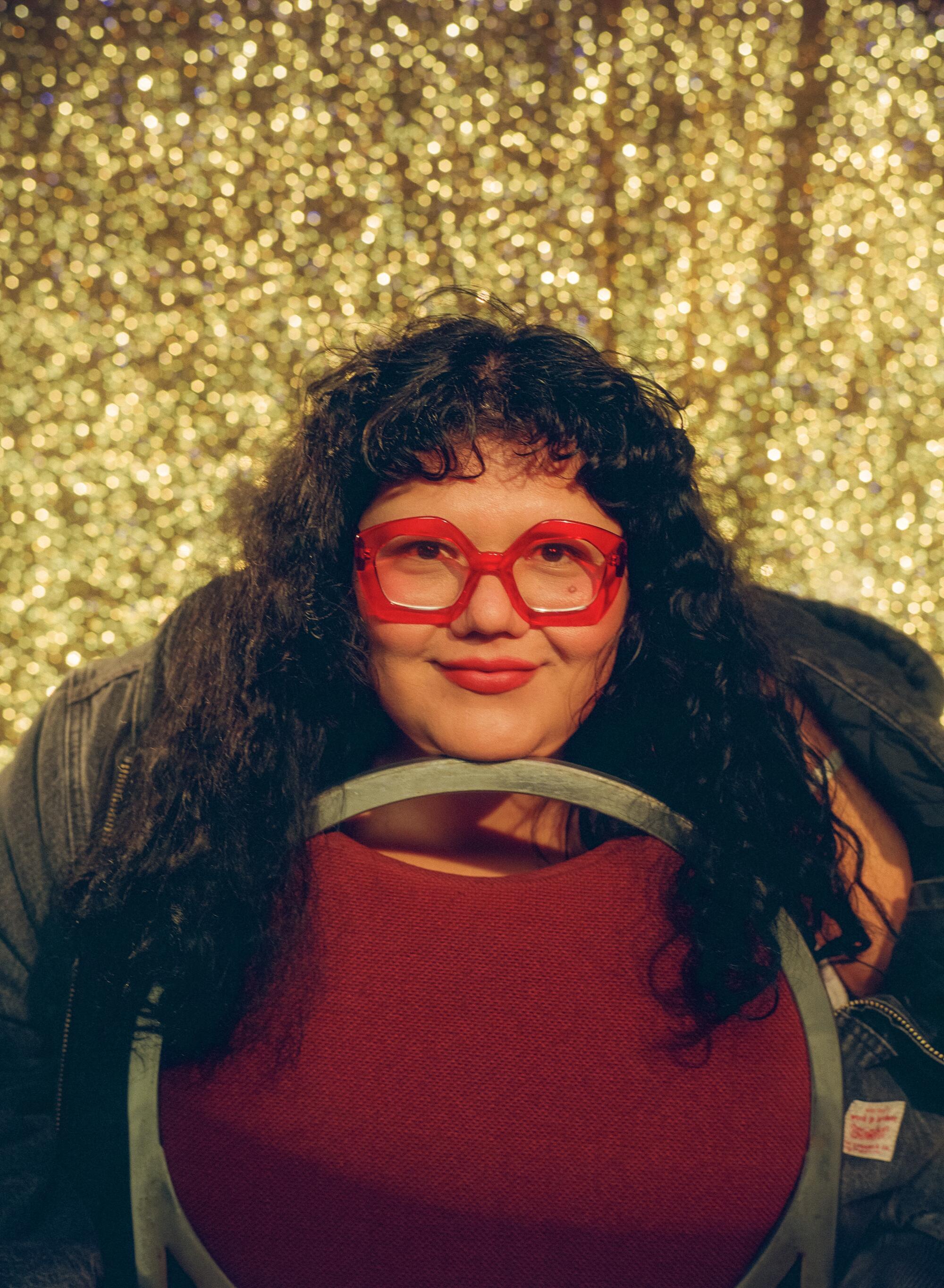 A woman in red glasses smiles before a golden curtain.