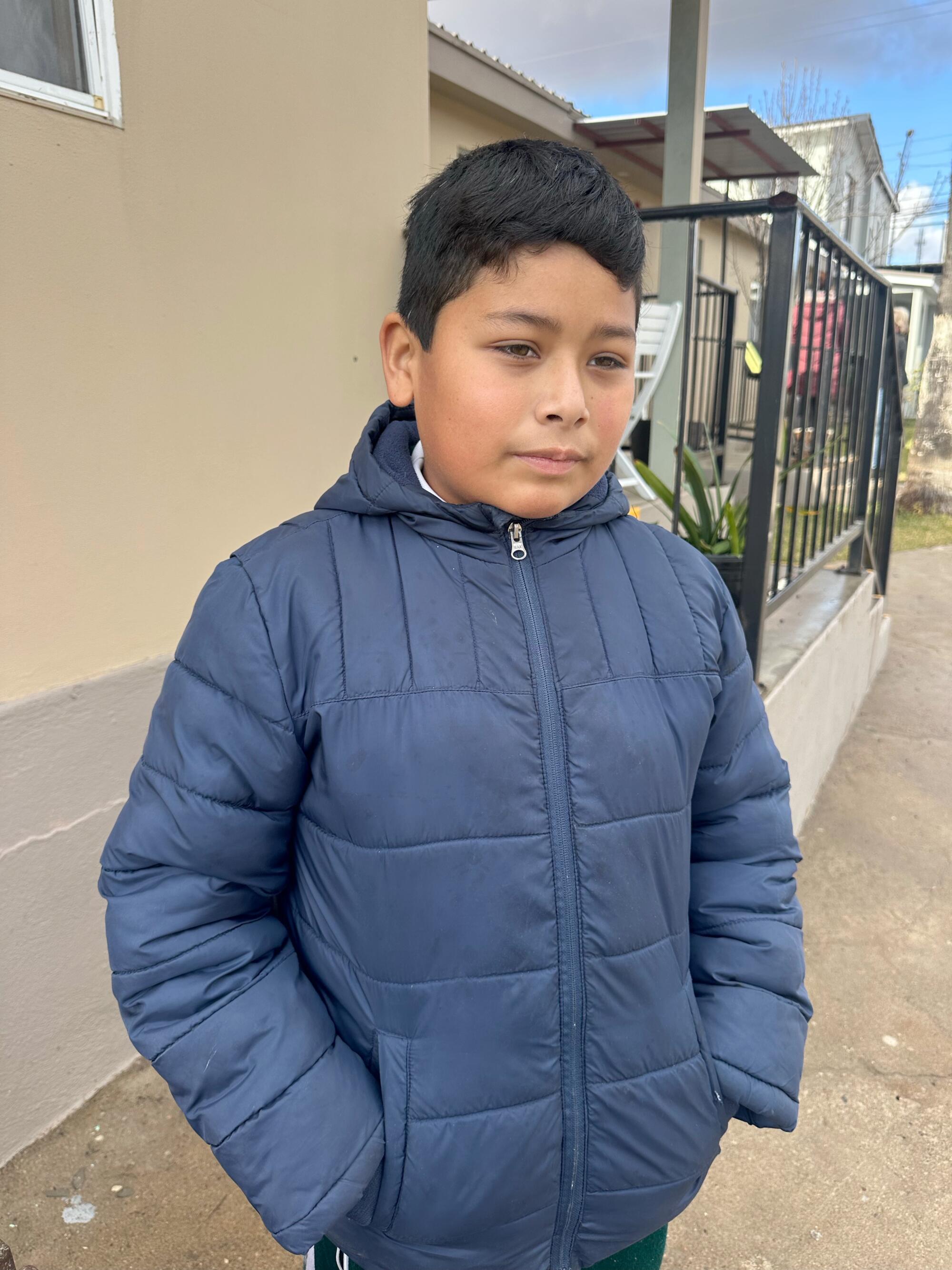 A boy in a blue jacket stands outside with his hands in his pockets.