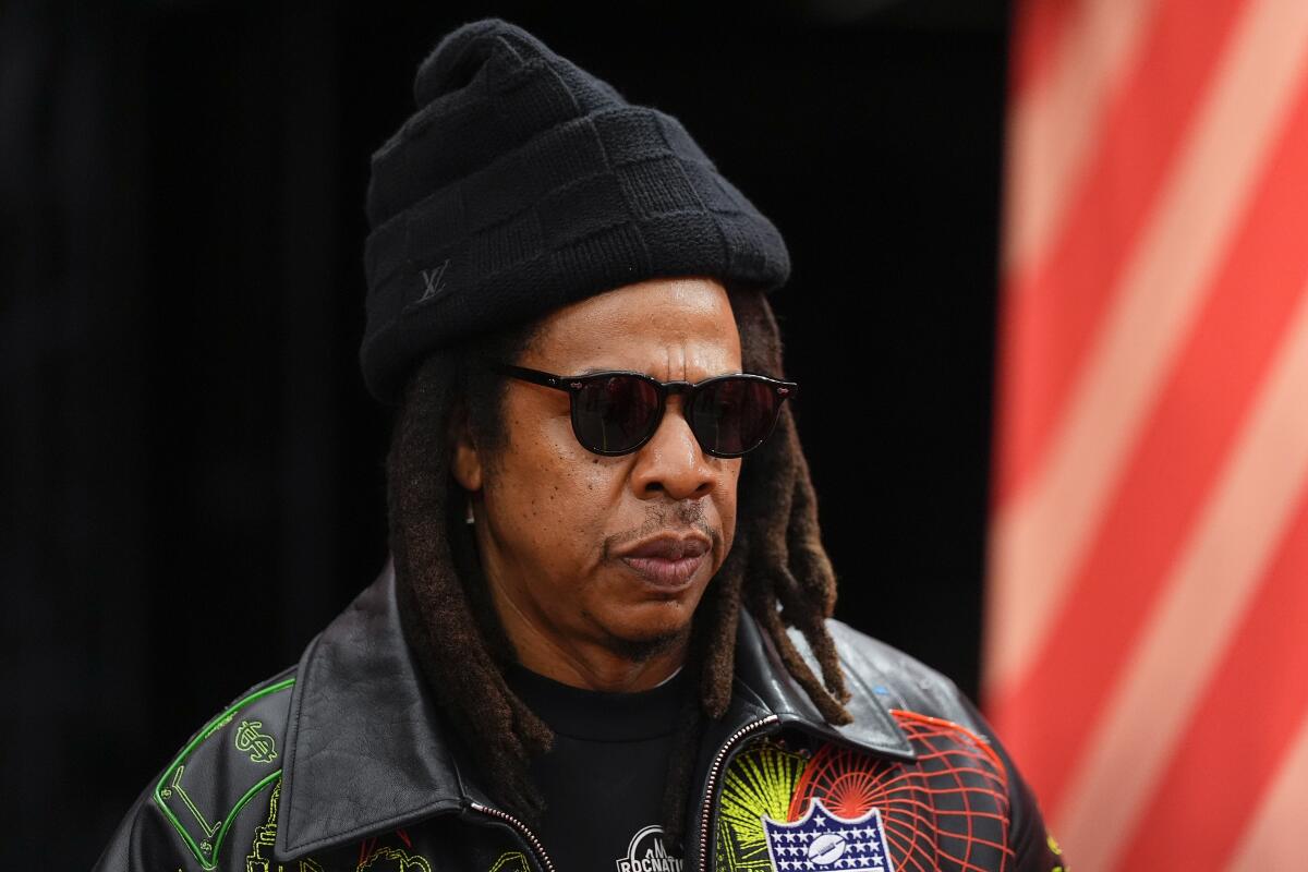 Jay-Z wears sunglasses and a black beanie in front of a dark background