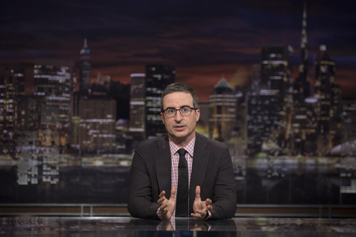 John Oliver sitting behind a desk with the New York skyline behind him.