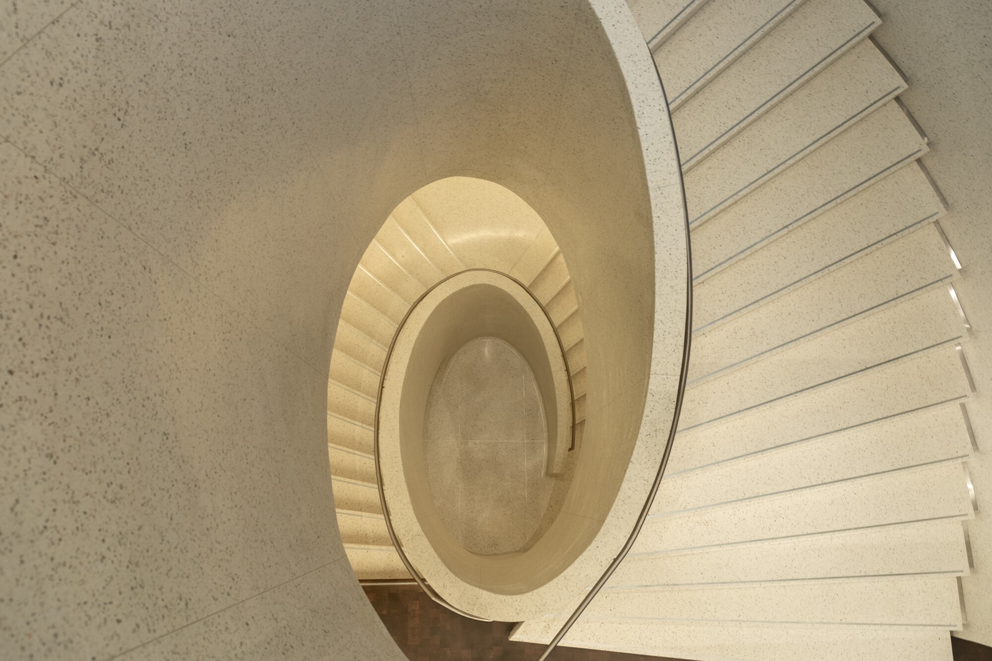 An overhead view shows an oval shaped staircase made from gleaming white terrazzo spiraling downwards