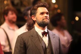 Chris Peluso smiling wearing brown suit and red bow tie while on stage, looking off at a crowd in front of cast