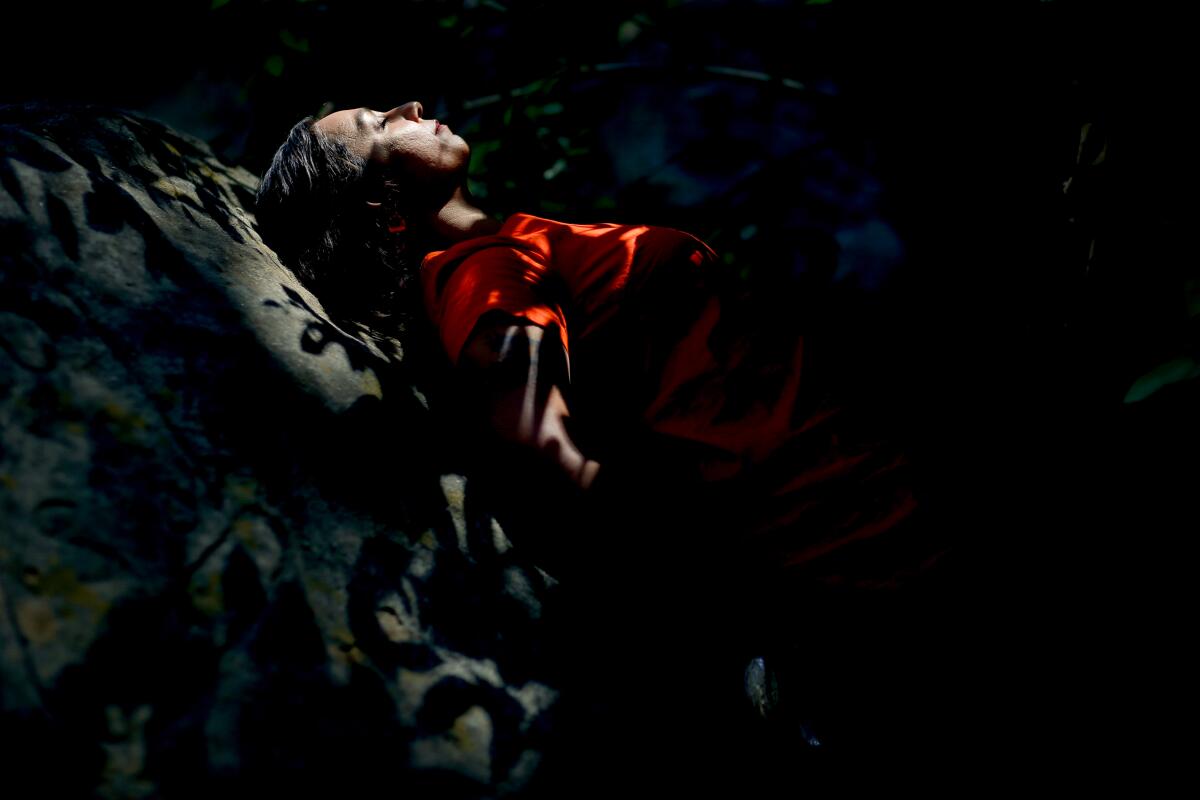 A woman in a bright orange dress leans back against a boulder with eyes closed and her face dappled by shadows.