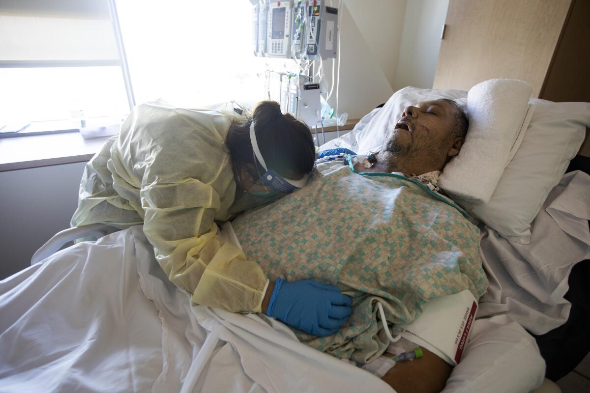 A woman in protective gear bends over the body of her father in a hospital bed