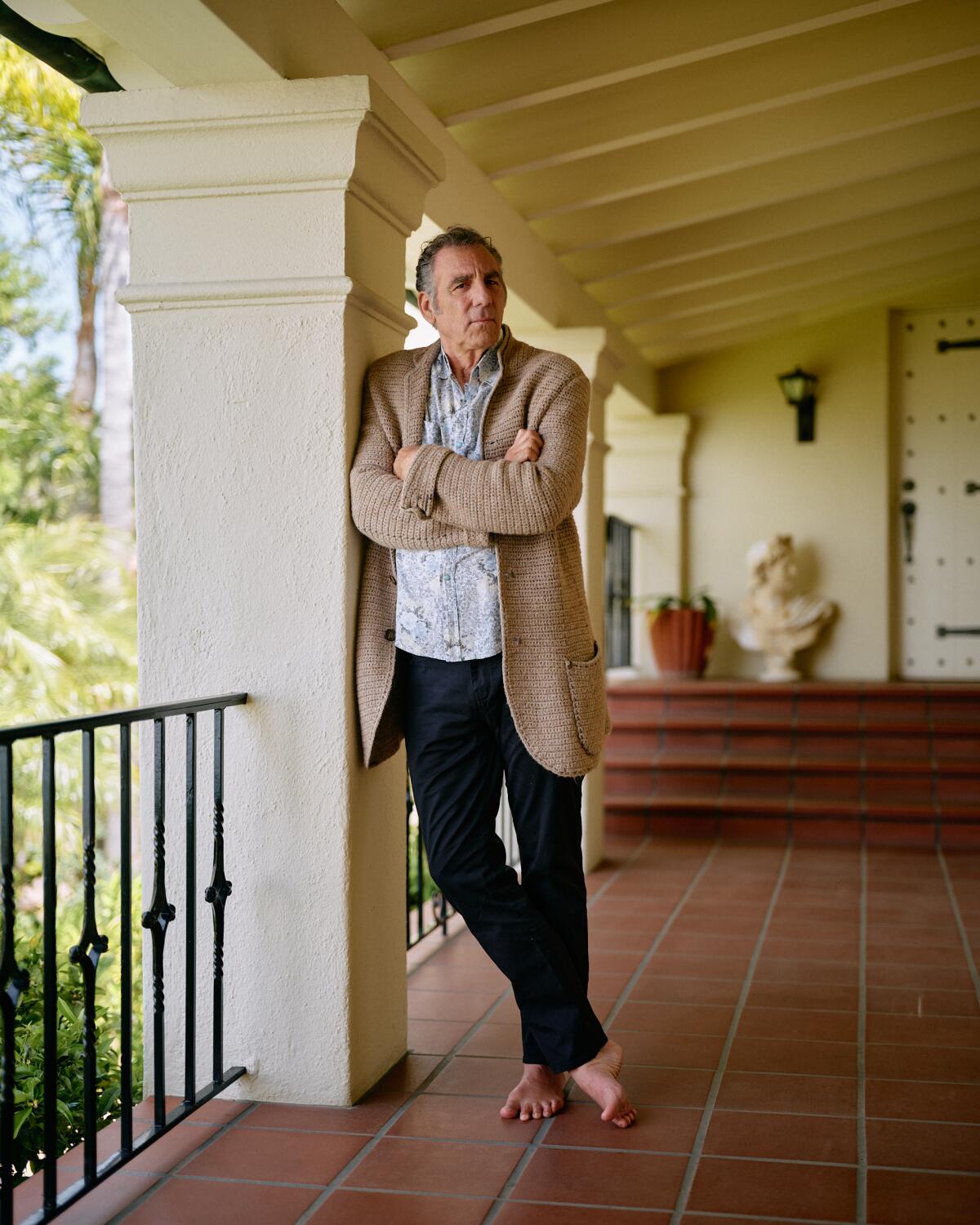 Michael Richards, barefoot and wearing a tan jacket and jeans, leans against a pillar.