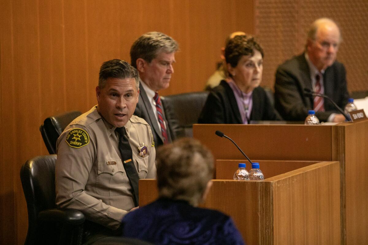 A man in a law enforcement uniform speaking as he sits at the front of a hearing room with several other people