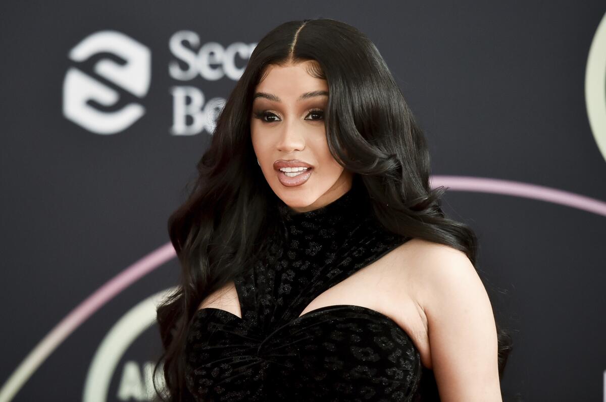 A woman with long hair wearing a black dress stands in front of a backdrop