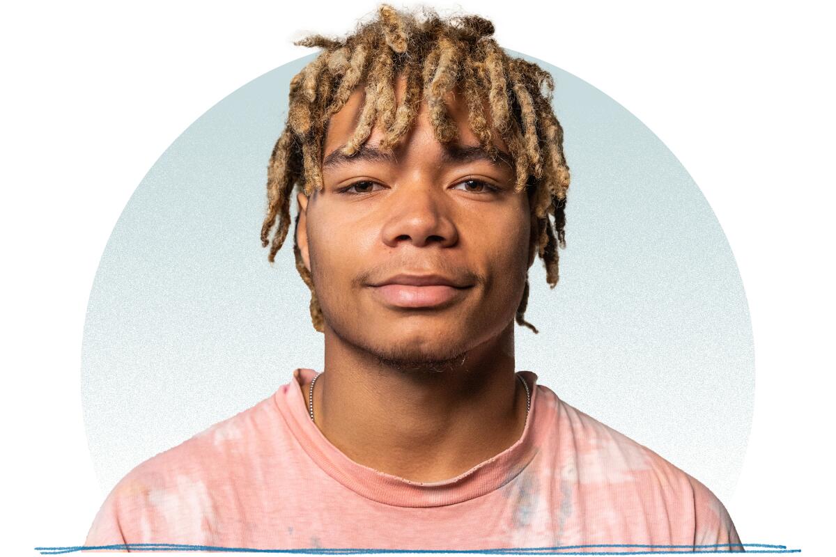 A young man with blond dreads and a pink shirt smiling for a portrait.