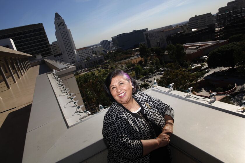 Gloria Molina poses for a portrait in sunlight, smiling. The LA City Hall is in the backgroung. 