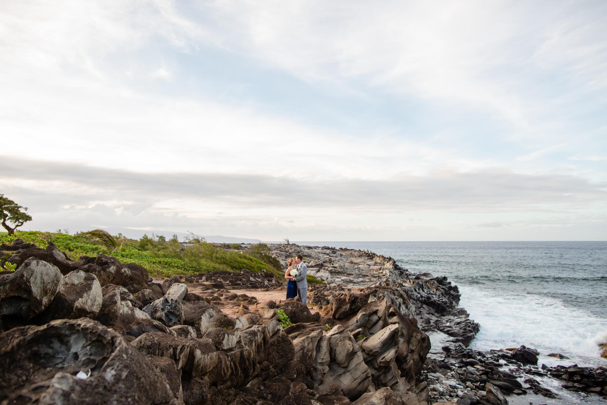 A couple stands on a rocky shoreline above the ocean
