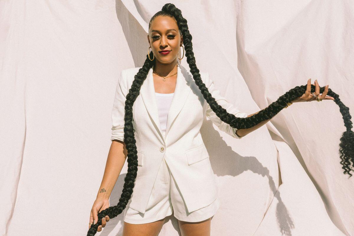 Tia Mowry strikes a pose in a white blazer while holding two long braids of hair.