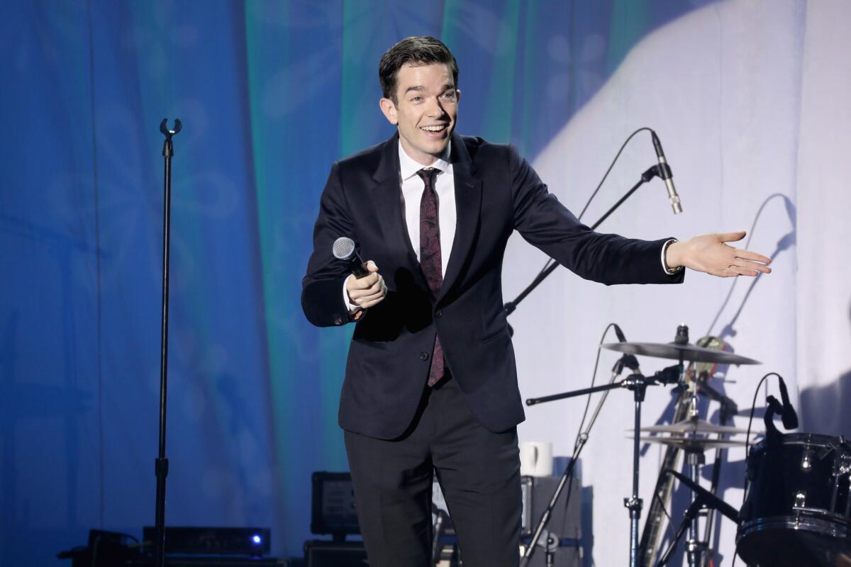 John Mulaney performing with a drum set in the background. 