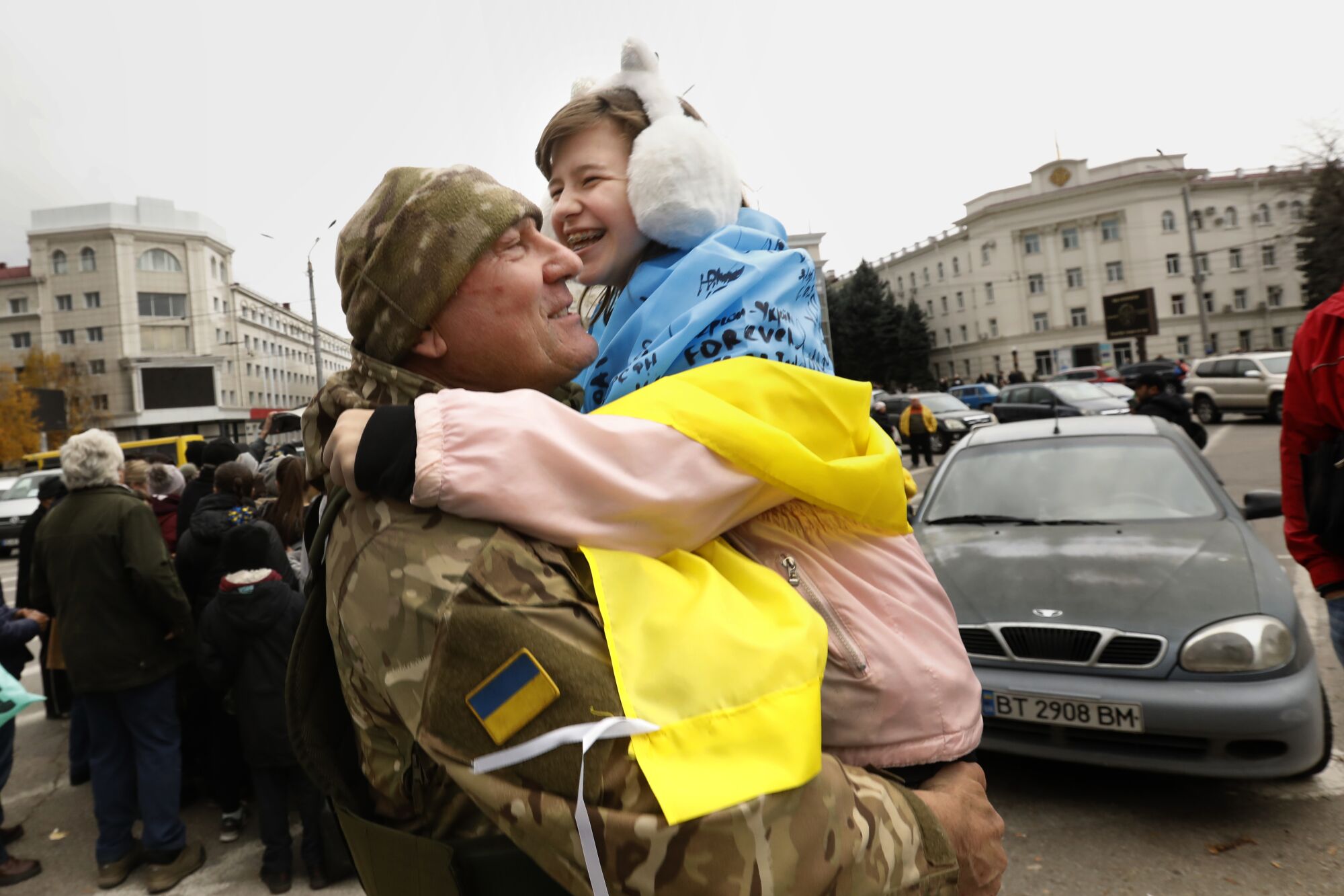 A man in military fatigues holds a smiling child wearing white earmuffs with a blue-and-yellow banner around her shoulders