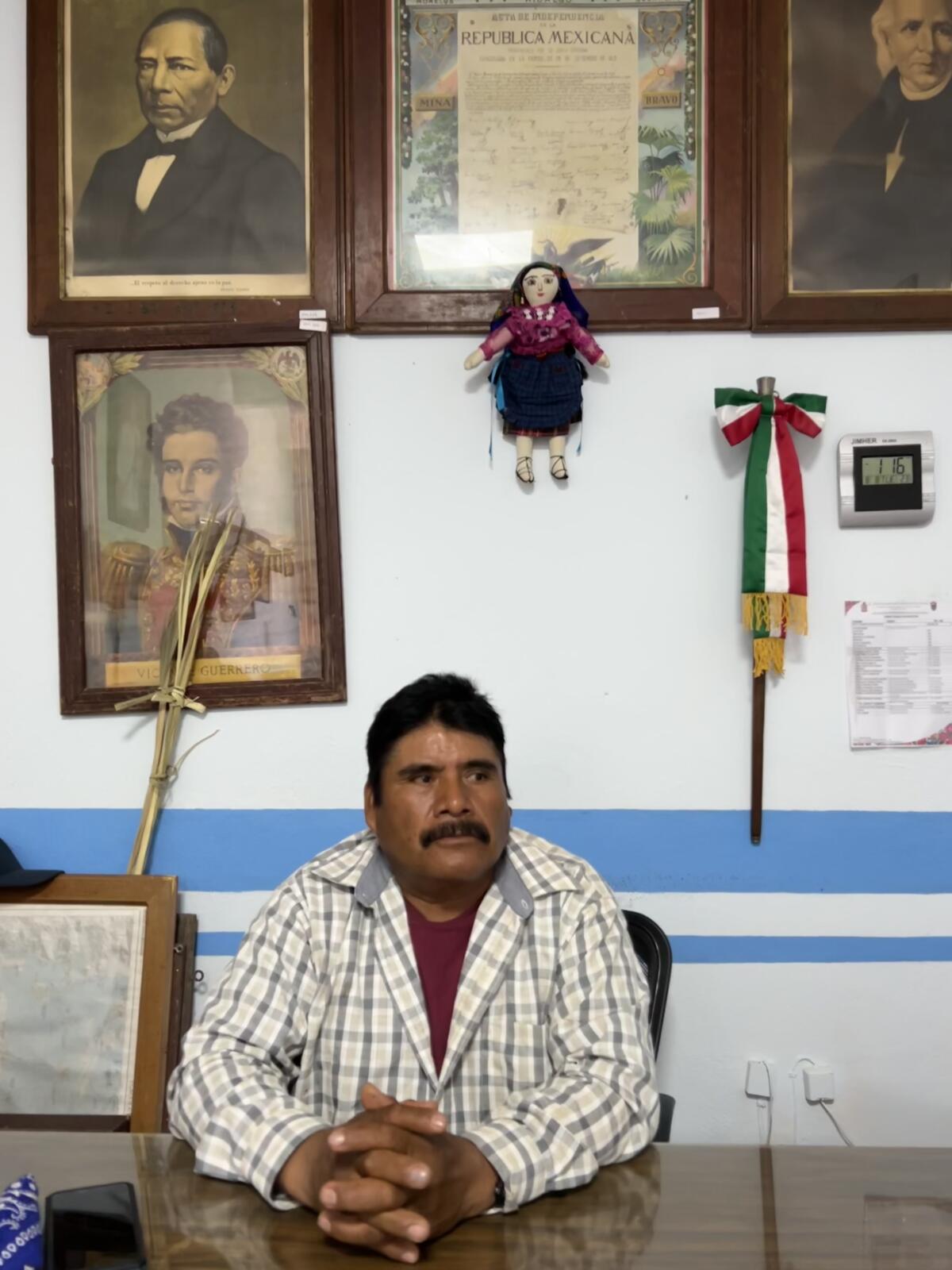 Мужчина сидит за столом перед стеной, на которой висят исторические мексиканские портреты. 