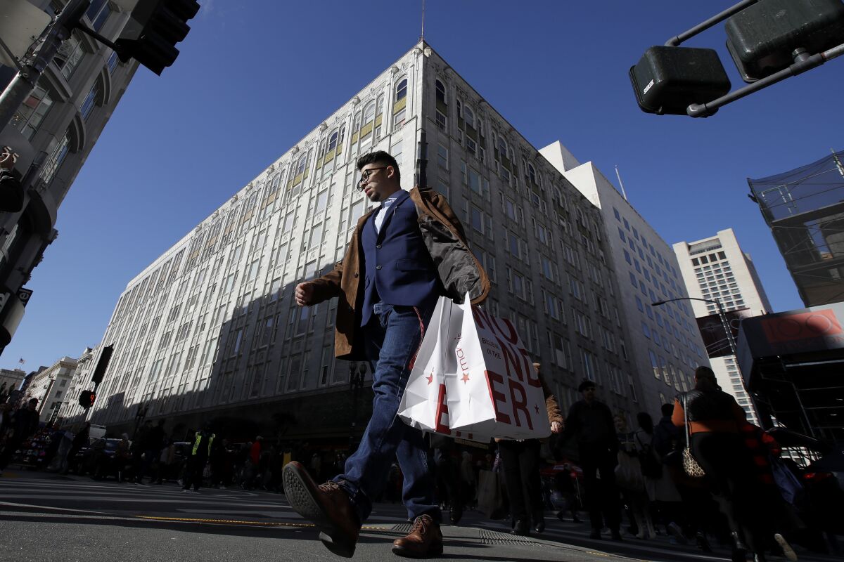 FILE - In this Nov. 29, 2019, file photo, man carries shopping bags across the street from a Macy's store in San Francisco. Macy's says it is closing 125 of its least productive stores and cutting 2,000 corporate jobs as the struggling department store tries to reinvent itself in the age of online shopping. The moves announced Tuesday, Feb. 4, 2020, come ahead of Macy's annual investor meeting where CEO Jeff Gennette is expected to unveil a three-year reinvention plan. (AP Photo/Jeff Chiu, File)