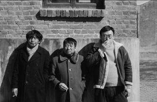 Three men wearing coats standing against a brick wall and smoking cigarettes