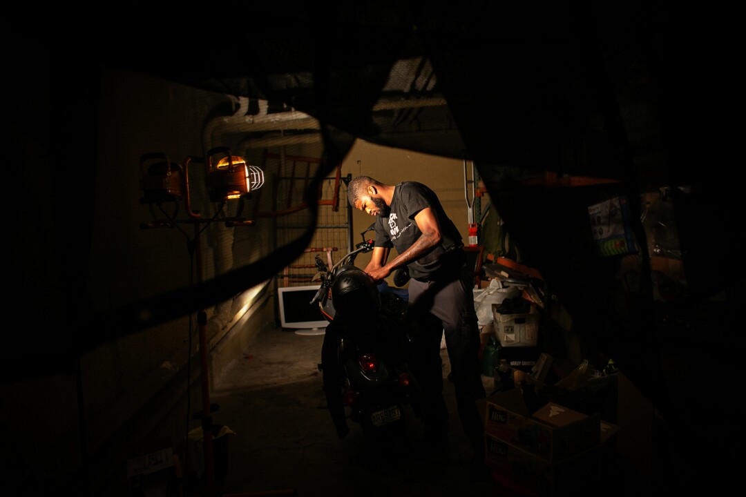 A man in a shadowed garage leans over a motorcycle.