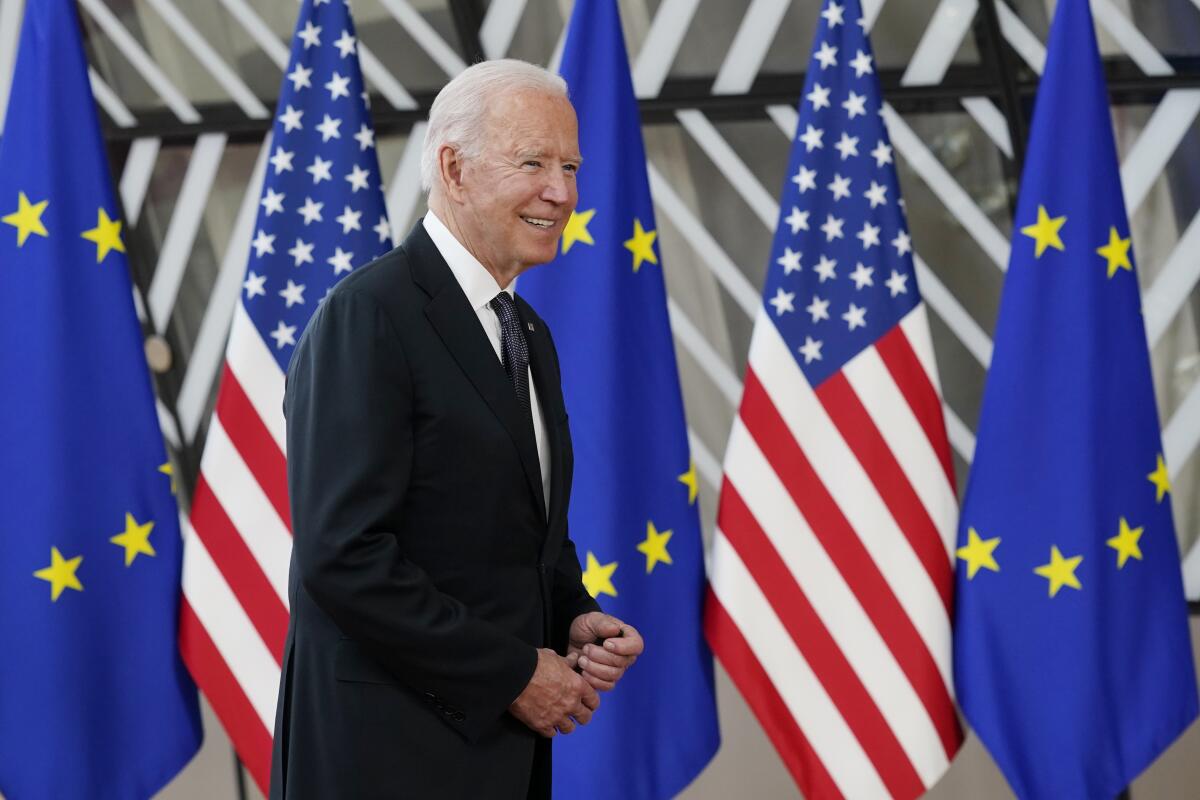 Biden, smiling, walks in front of U.S. and EU flags.