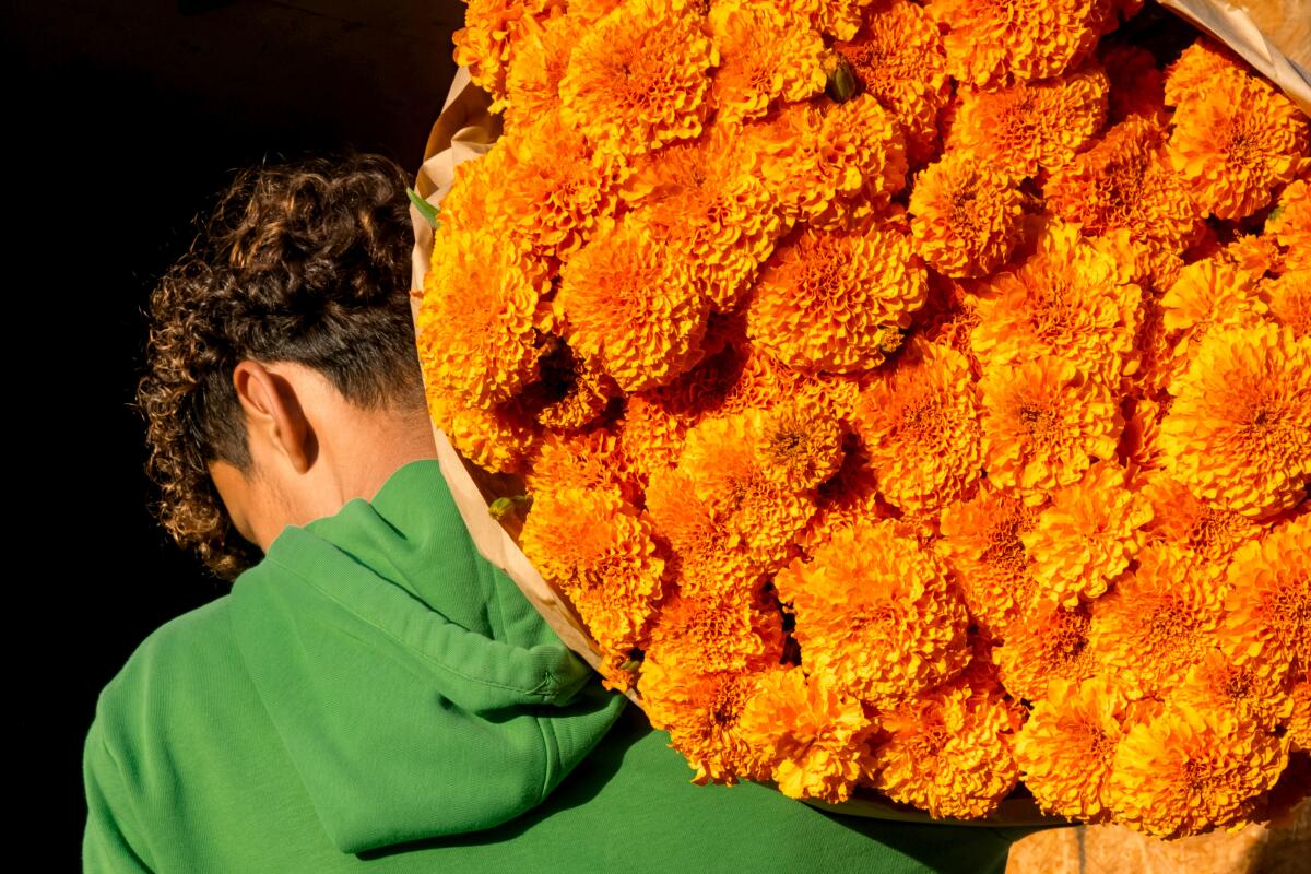 A person in a green hoodie, seen from behind, with a large bundle of orange marigolds 