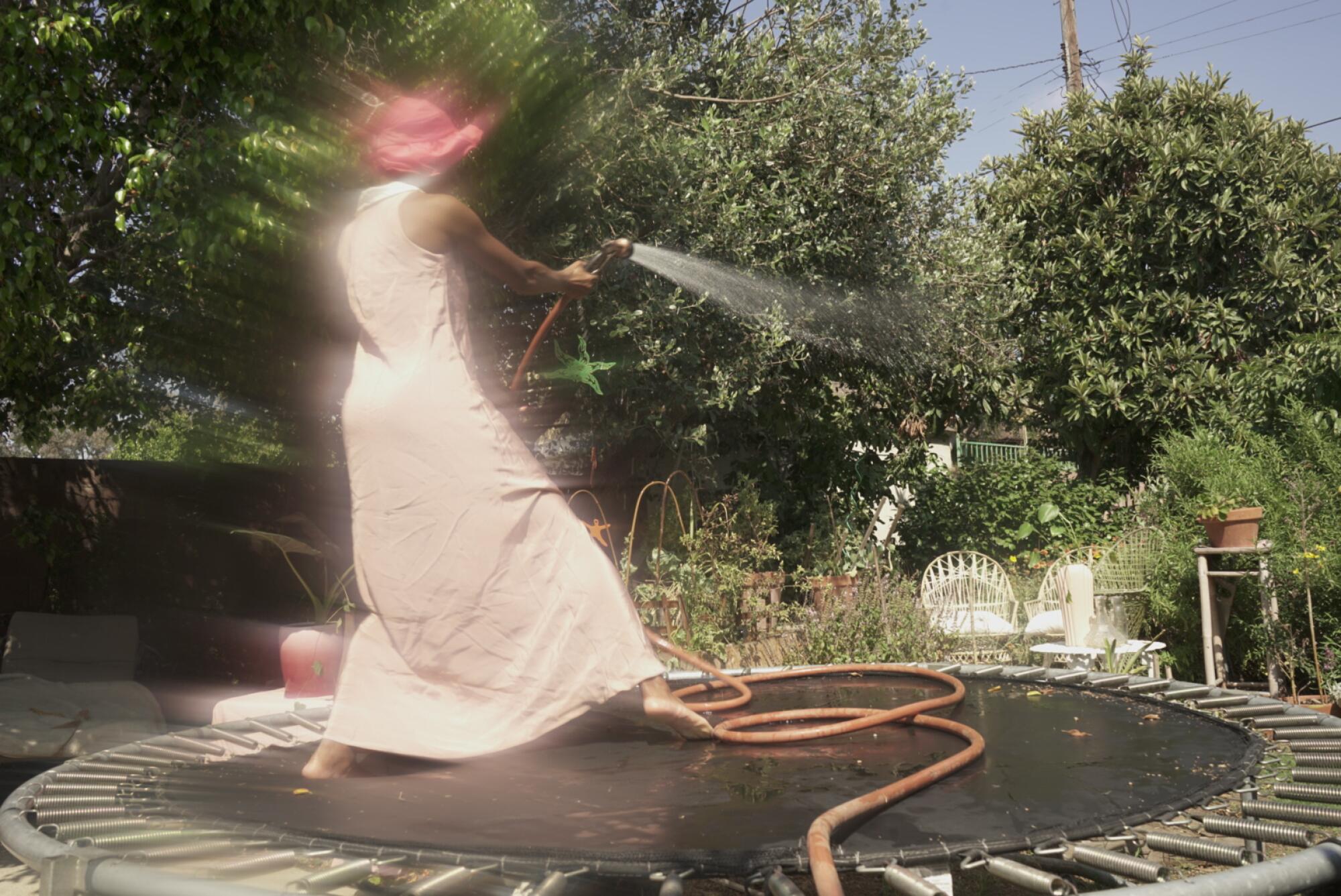 A woman in a long light-colored dress and hat stands on a trampoline spraying plants with a hose.