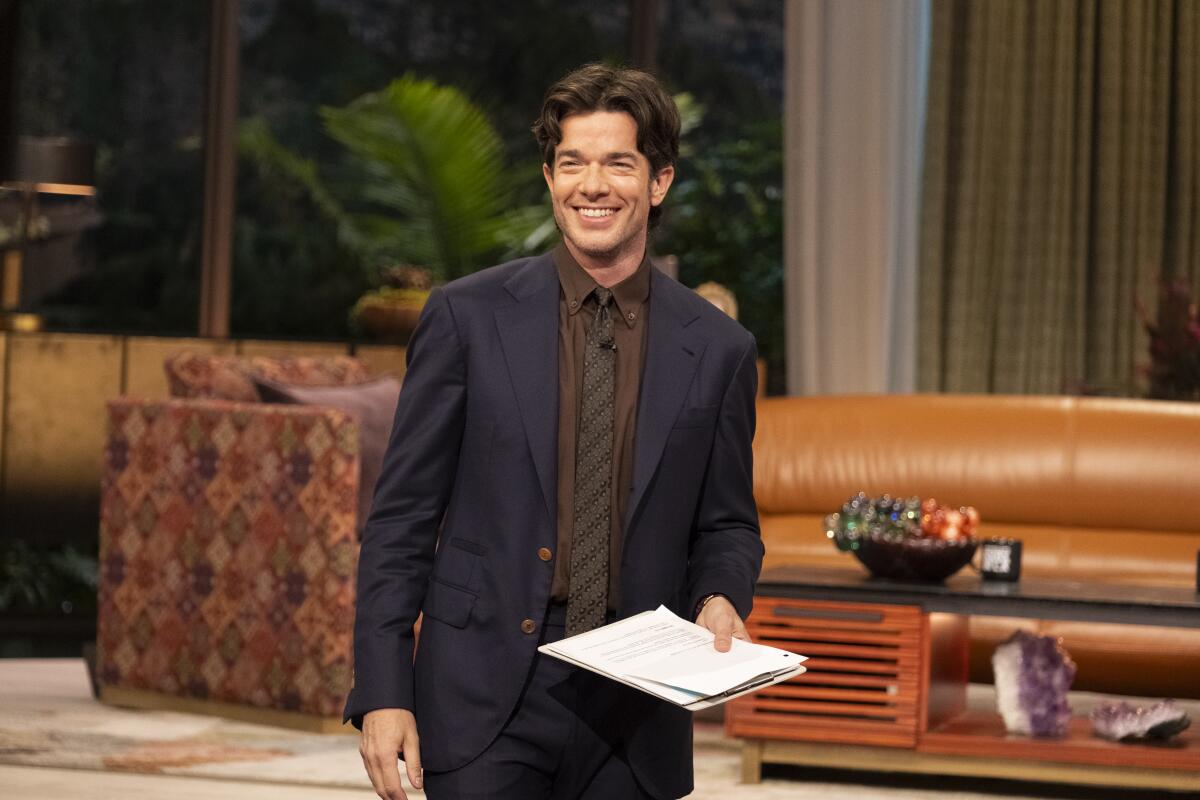John Mulaney standing on the set of his show with papers in hand.