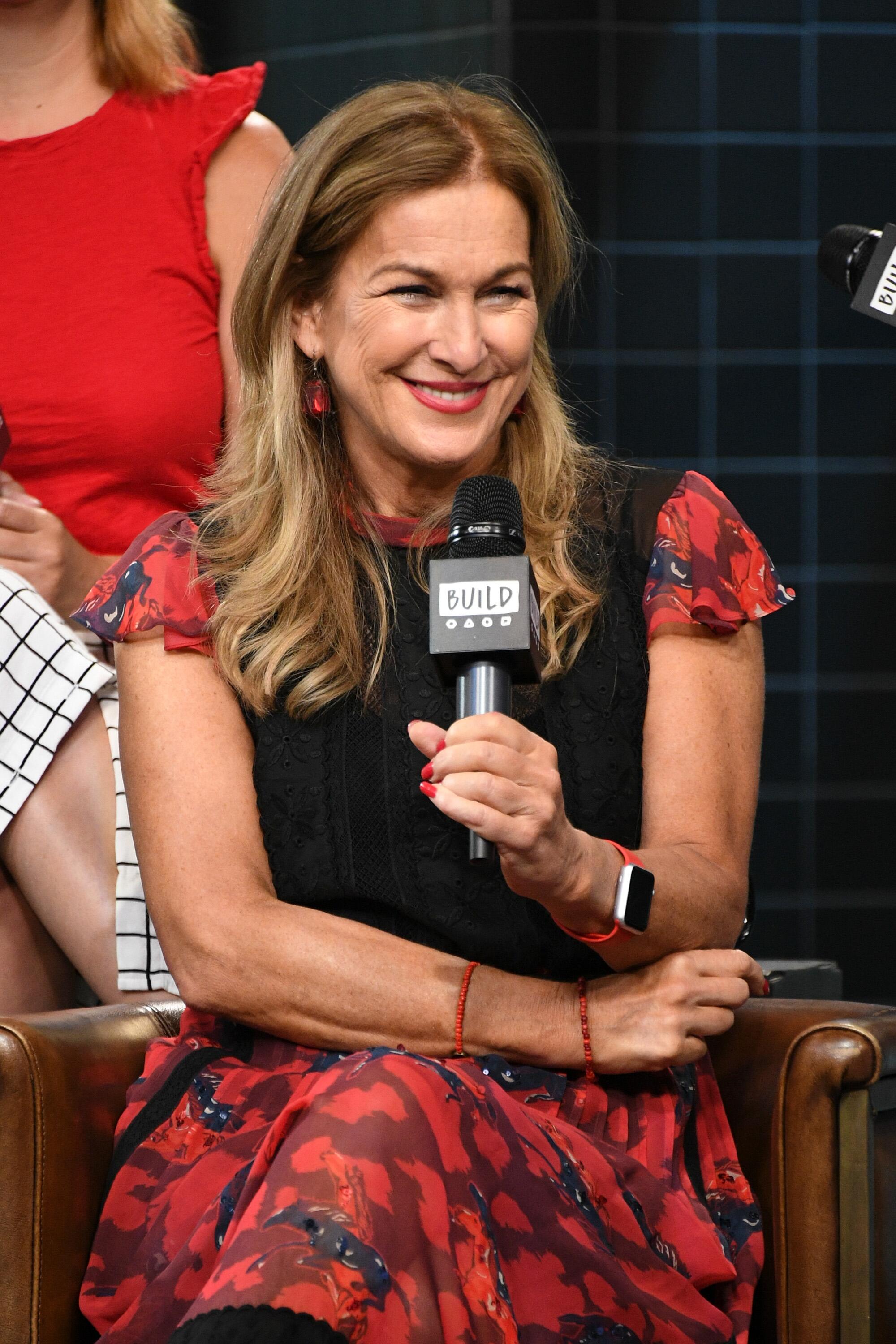 A woman in a red and black outfit, seated, smiling and holding a microphone